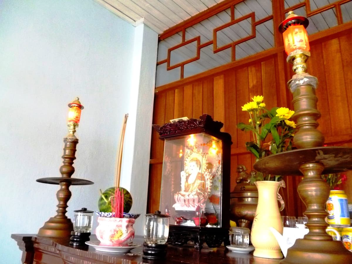 Bàn thờ gỗ có bát hương cắm nhang, hai chân đèn đồng, lọ hoa cúc vàng, quả dưa hấu và khám kính có tượng Phật ở giữa.