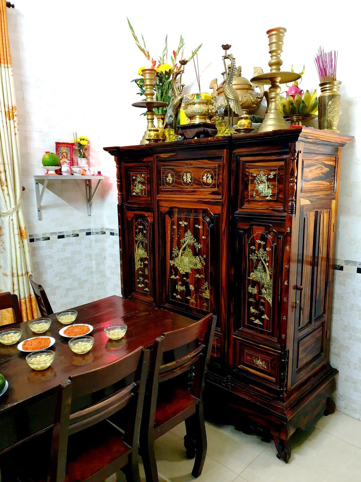 Tủ thờ gỗ khảm xà cừ, trên nóc có lư hương, chân nến, đôi hạc đồng, lọ hoa lay ơn và mâm chuối có thanh long; bàn bên cạnh bày các chén chè và đĩa xôi gấc.