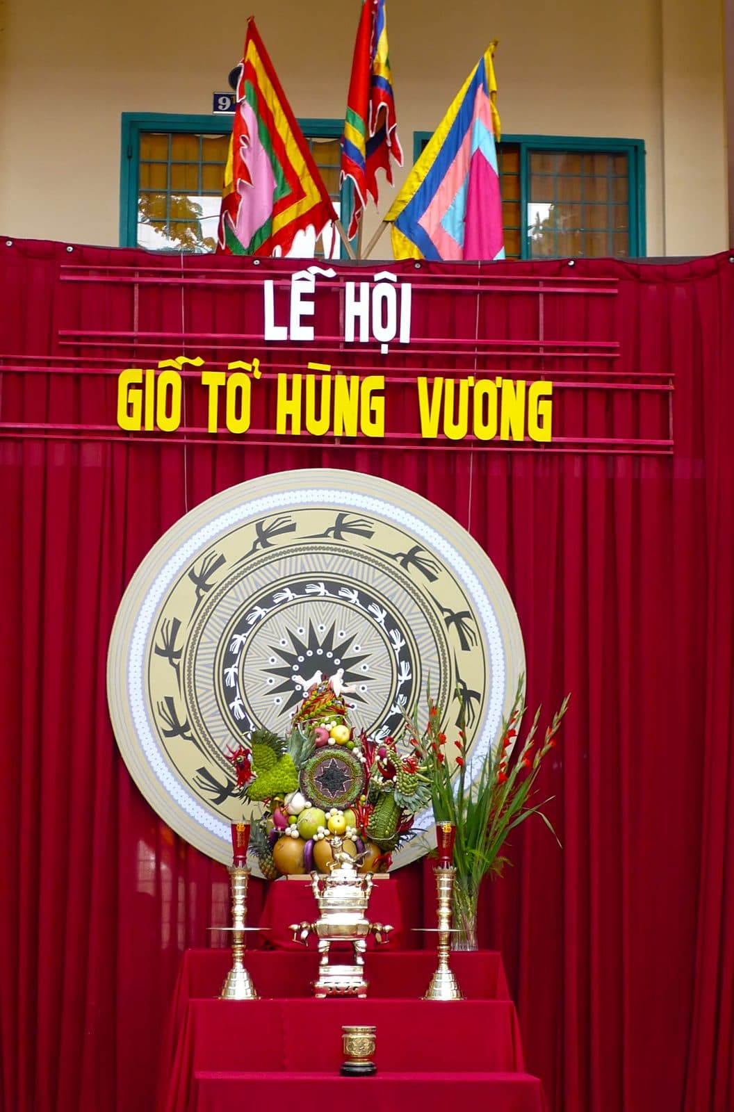 A three-tier altar draped in red with a brass incense burner, two candlesticks, a vase of gladioli and a tall arranged fruit tray; behind it a large bronze-drum face, the words Lễ hội Giỗ Tổ Hùng Vương and five-colour flags.
