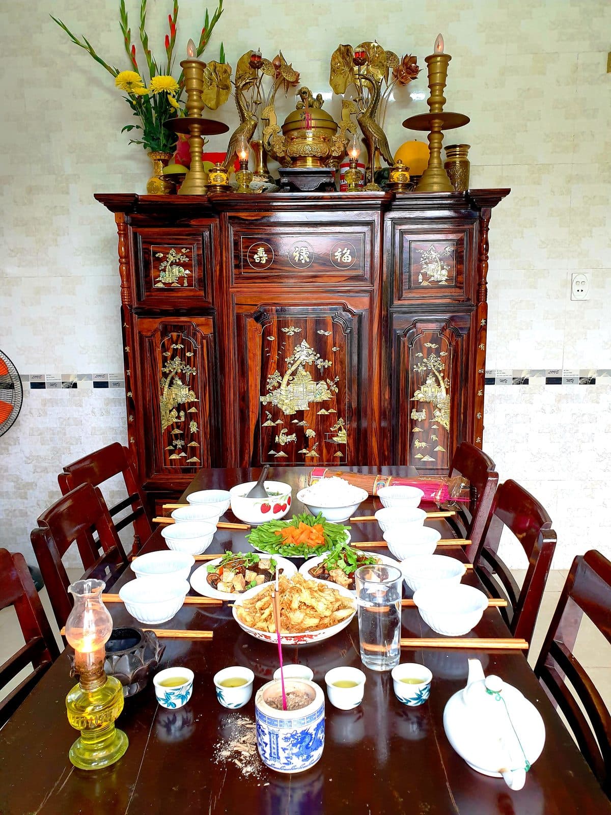 Bàn gỗ trước tủ thờ bày các món xào, đậu phụ rán, đậu que xào cà rốt, bát cơm, hai hàng bát đũa, đèn dầu, bát hương, chén trà và ấm trà.