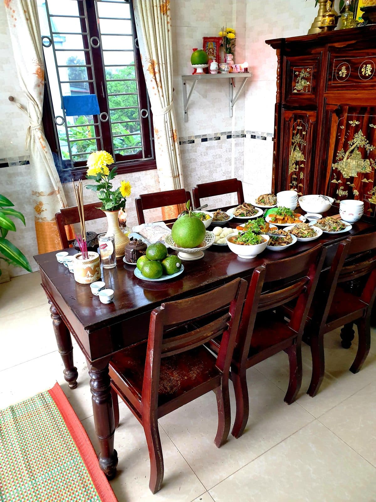 Bàn gỗ dài bày bát hương, lọ hoa cúc vàng, đĩa cam, quả bưởi, đĩa bánh bao và nhiều đĩa thức ăn, cạnh tủ thờ khảm xà cừ.