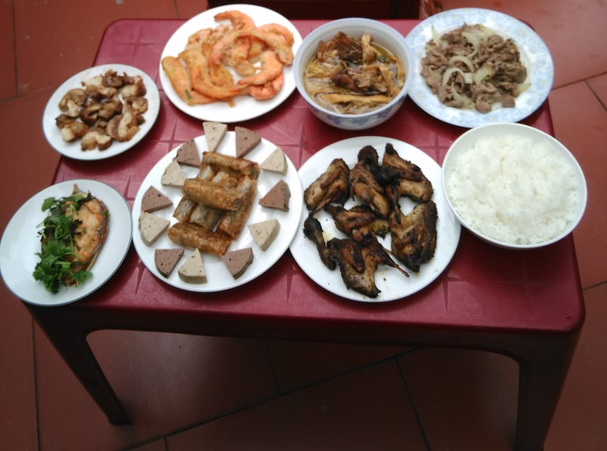 Bàn nhựa đỏ bày tôm chiên, chả, nem rán, cánh gà nướng, thịt bò xào hành, cá, bát canh măng và bát cơm.