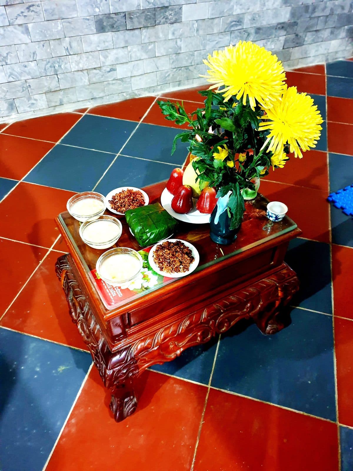 Bàn gỗ thấp chạm trổ bày lọ hoa cúc vàng, đĩa mận và quả bưởi, bánh chưng, các bát chè, đĩa xôi và chén nước.