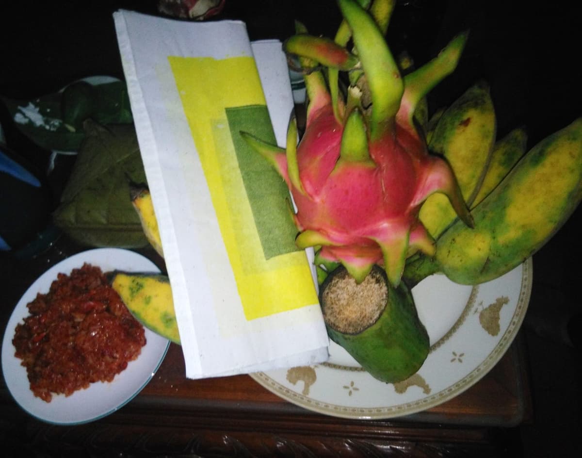 Close-up of a dish of dragon fruit and bananas, a plate of red sticky rice, leaf-wrapped cakes and a pile of votive paper on a table.