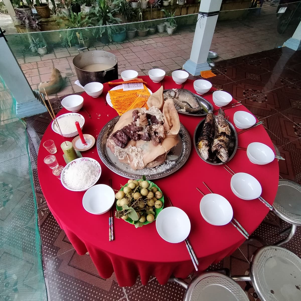 Bàn tròn phủ khăn đỏ với đĩa thịt luộc lớn ở giữa, hai đĩa cá nướng, đĩa nhãn, hai bát gạo, chén rượu, nến đỏ, nhang, giấy tiền vàng và một vòng bát đũa.
