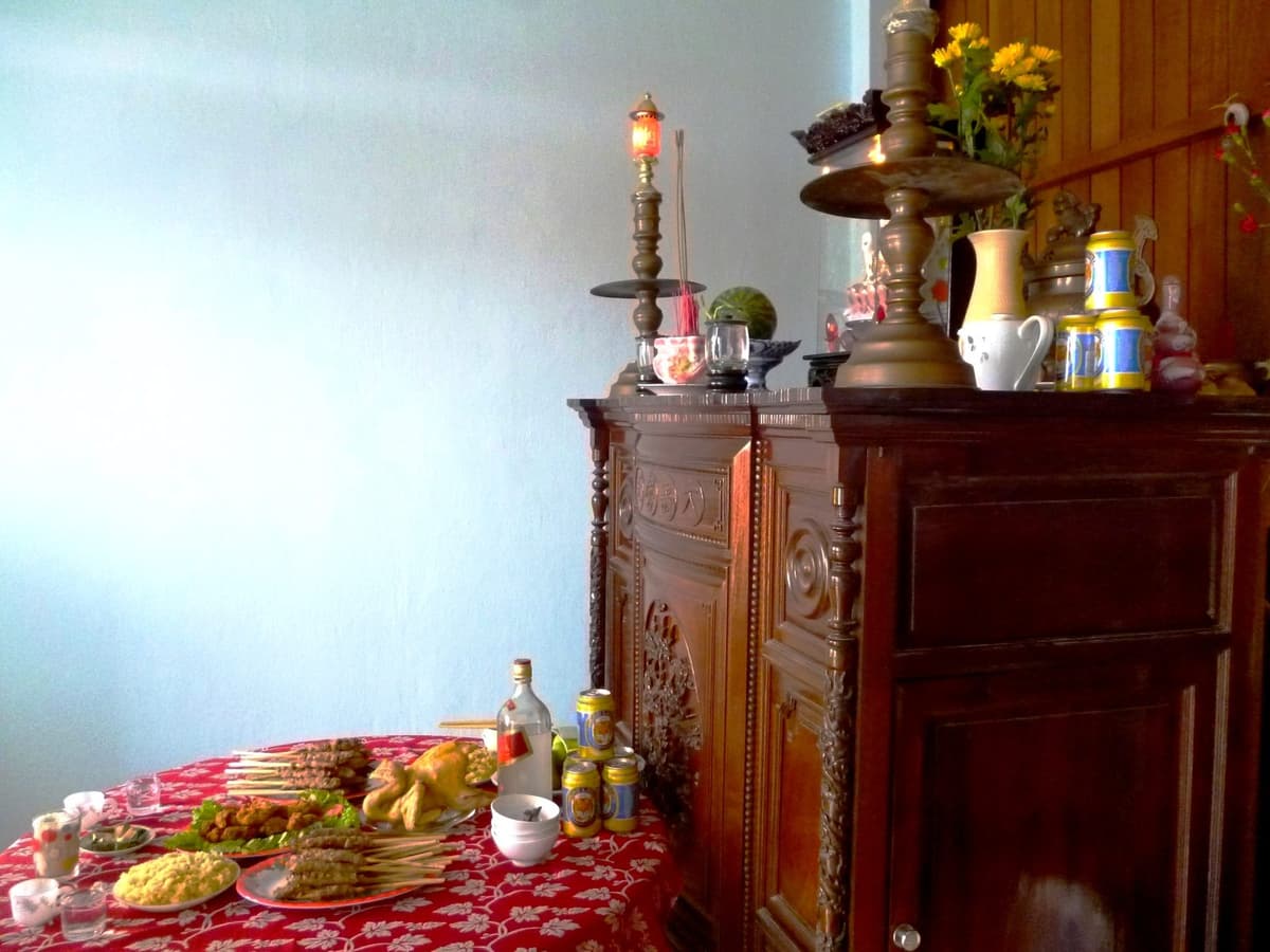 Bàn phủ khăn đỏ hoa trắng bày gà luộc, nem nướng xiên que, món chiên, xôi vàng, chai rượu, lon nước và chồng bát, cạnh tủ thờ gỗ có chân đèn đồng và lọ hoa cúc.