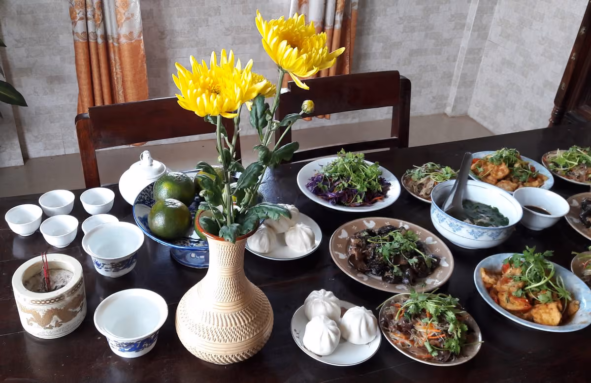 Bàn gỗ bày bát hương, lọ hoa cúc vàng, đĩa cam, hai đĩa bánh bao, chén trà, nhiều đĩa món xào và một bát canh.