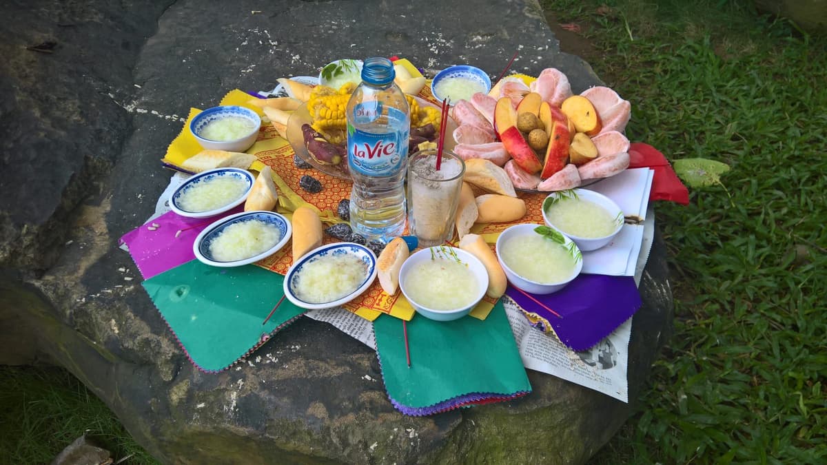 Trên tảng đá bày nhiều bát cháo trắng, bánh mì, ngô luộc, khoai lang, đĩa bưởi và táo, chai nước, cốc gạo cắm nhang và giấy cúng nhiều màu.