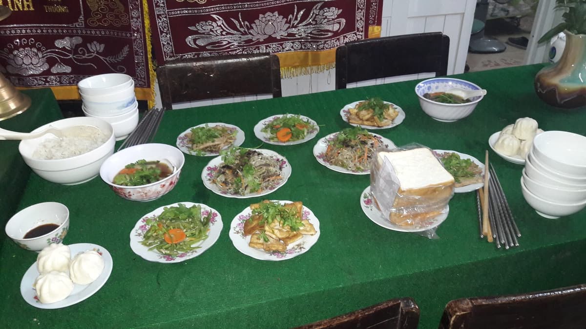A table covered in green cloth with many vegetarian dishes, steamed buns, a wrapped loaf of bread, a bowl of rice, a bowl of soup and stacks of bowls and chopsticks.