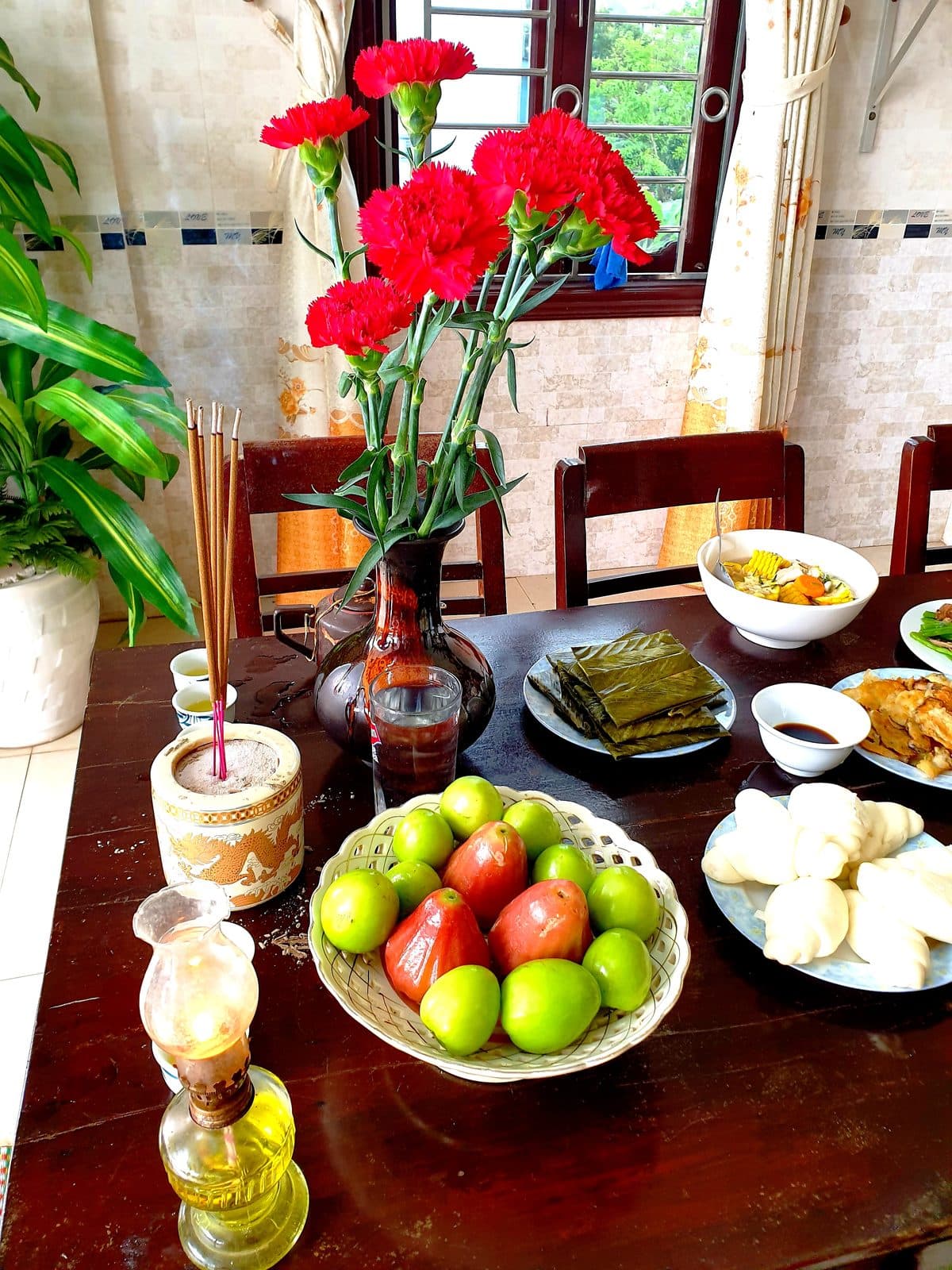 A wooden table with an incense pot, an oil lamp, a vase of red carnations, a dish of jujubes and rose apples, a plate of steamed buns, leaf-wrapped cakes and vegetarian dishes.