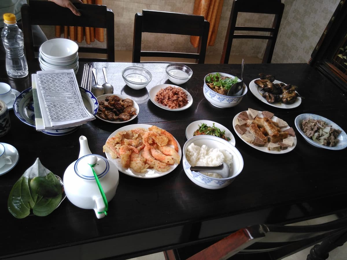 A dark wooden table with fried prawns, slices of pork roll, fried spring rolls, meat dishes, salad, a bowl of rice, a teapot, a stack of bowls, a pile of votive paper and betel leaves.