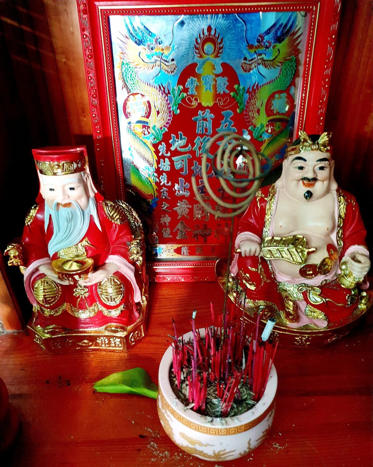 Tượng sứ Thần Tài và Ông Địa hai bên bát hương cắm đầy chân nhang, nhang vòng treo phía trên, sau là bài vị đỏ in chữ Hán và hình rồng.