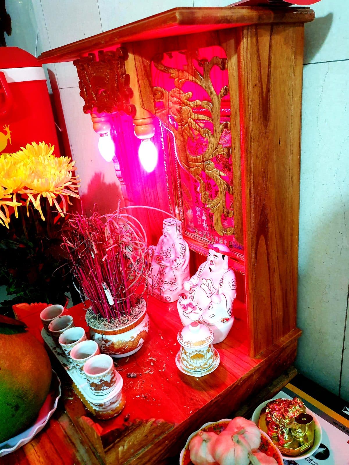 Khám thờ gỗ đặt dưới đất, đèn đỏ chiếu hai tượng sứ trắng, bát hương, hàng chén nước, hũ nhỏ có nắp, tượng cóc vàng, quả bưởi và hoa cúc vàng.