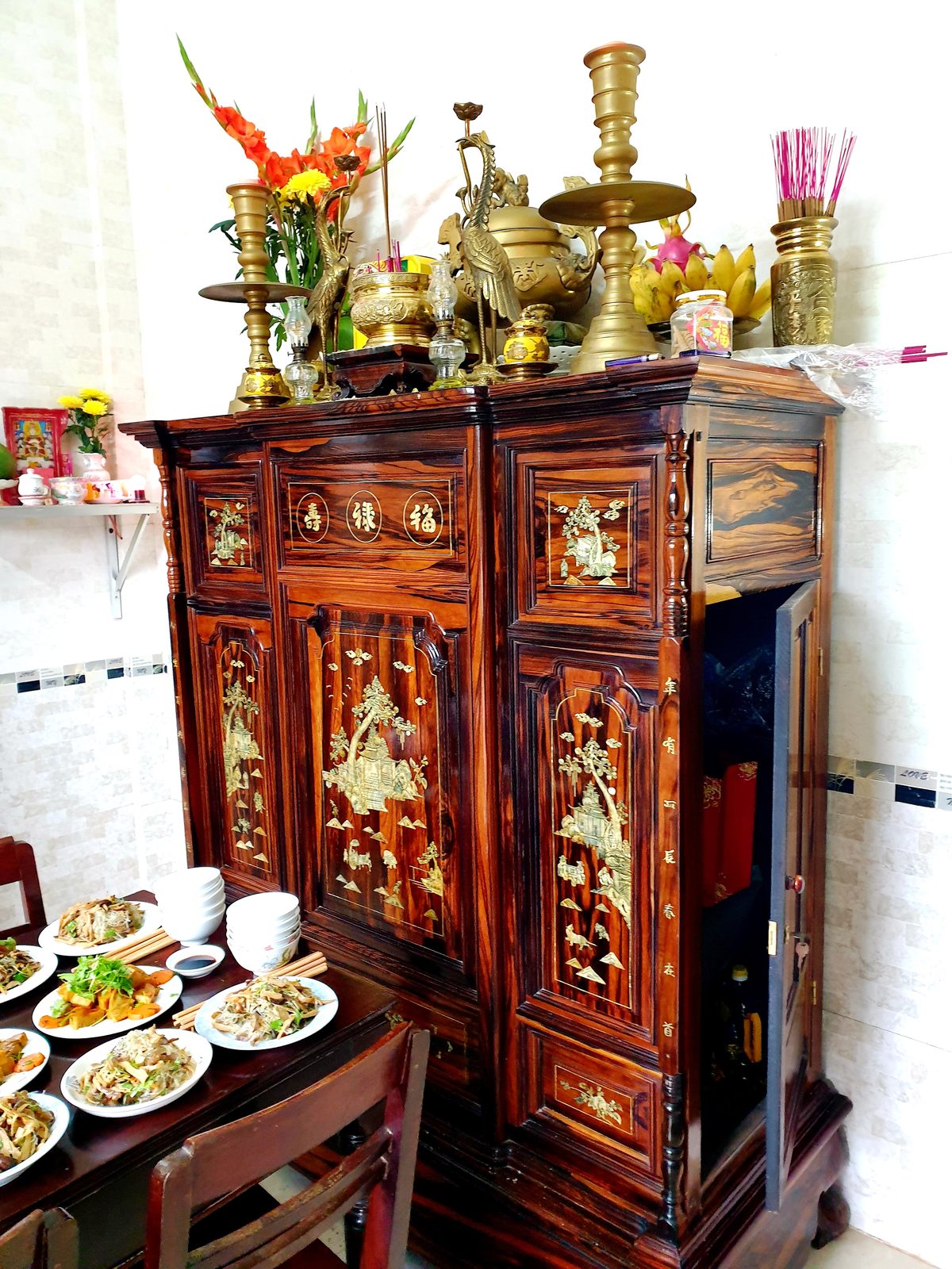 Bàn thờ gia tiên bằng gỗ chạm khắc với bát hương, chân đèn đồng, hoa và trái cây.